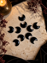 Load image into Gallery viewer, Ten polished black obsidian crescent moon carvings arranged on cream stone with candlelight and dried botanicals