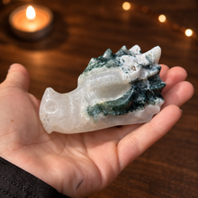 Load image into Gallery viewer, Moss agate dragon head carving held in hand with dark wood and candlelight background