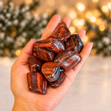 Load image into Gallery viewer, Red tiger’s eye tumbled stones held in hand against ivory, evergreen and golden holiday lights