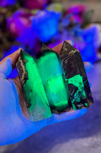 Load image into Gallery viewer, A hand holding two green Fluorescent Volcano Agate crystals under UV light, showcasing their glowing green color against a blurred floral background.