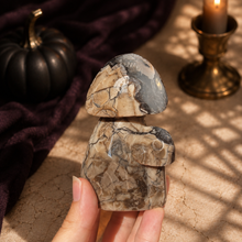 Load image into Gallery viewer, Cream gray and black Volcano Agate mushroom carving held beside a black pumpkin and candle
