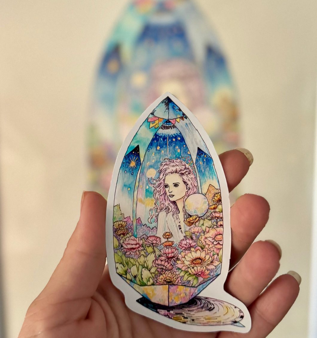 Hand holding a colorful teardrop-shaped sticker with a fantasy design of a woman and nature elements.