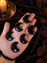 Load image into Gallery viewer, Six polished black obsidian crescent moon carvings held in a hand against black velvet with warm candlelight
