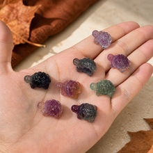 Load image into Gallery viewer, Seven mini fluorite turtle carvings resting in Eva's open hand against pale stone with restrained rust leaves and autumn tones
