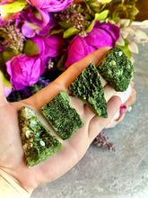Load image into Gallery viewer, A hand holding several green epidote specimens with a background of purple flowers.
