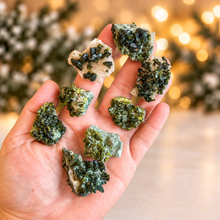 Load image into Gallery viewer, Raw green epidote specimens held in hand with softly blurred evergreen and golden Christmas lights
