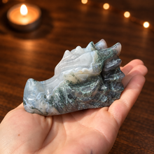 Load image into Gallery viewer, Moss agate dragon head carving held in hand with dark wood and candlelight background
