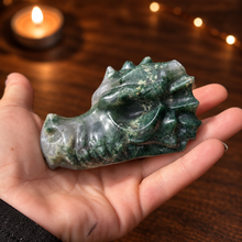 Load image into Gallery viewer, Moss agate dragon head carving held in hand with dark wood and candlelight background
