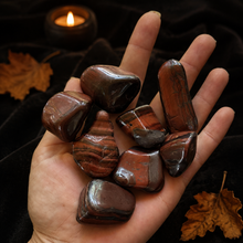 Load image into Gallery viewer, Red Tiger's Eye tumbled stones held in hand on black velvet with autumn leaves and candlelight
