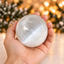 Load image into Gallery viewer, White selenite sphere cradled in a hand with blurred evergreen and golden holiday lights
