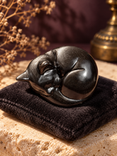 Load image into Gallery viewer, Single curled silver sheen obsidian cat carving displayed on charcoal velvet and limestone
